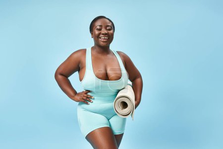 Lächelnde plus size afrikanisch-amerikanische Frau steht mit Fitnessmatte auf blauem Hintergrund, Körper positiv