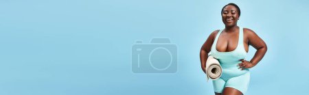 fröhliche plus size afrikanisch-amerikanische Frau mit Fitnessmatte auf blauem Hintergrund, Sportbanner