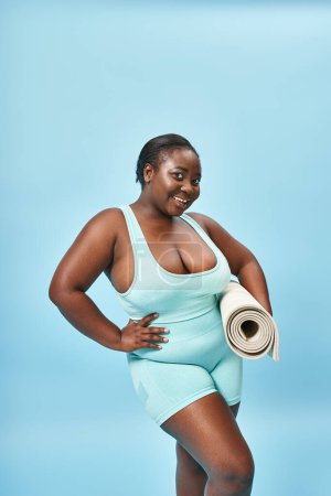 happy plus size african american woman standing with fitness mat on blue backdrop, sport