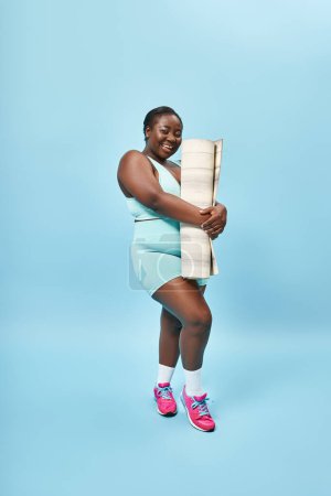 Lächelnde plus size afrikanisch-amerikanische Sportlerin mit Fitnessmatte auf blauem Hintergrund, Sport