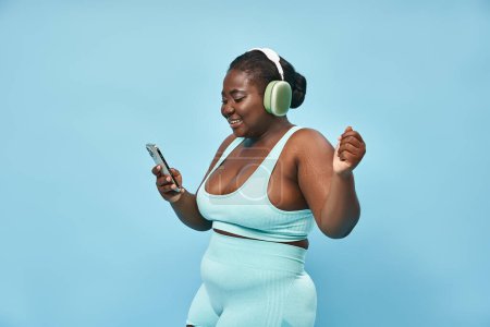 plus size afrikanisch-amerikanische Frau, die Musik im Kopfhörer hört und Smartphone in der Hand hält, blauer Hintergrund