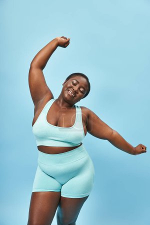cheerful plus size african american woman in active wear stretching joyfully on blue background