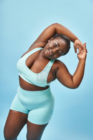 fröhliche plus size afrikanisch-amerikanische Frau in aktiver Kleidung Stretching auf blauem Hintergrund, Körper positiv