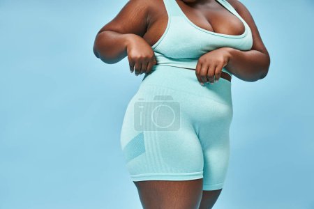 Detaillierte Ausschnittansicht der Plus-Size-Sportlerin in Radlerhosen auf blauem Hintergrund, Körper positiv