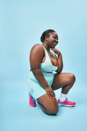 volle Länge glücklich plus Größe Sportlerin in Radlerhosen sitzt auf blauem Hintergrund, Körper positiv