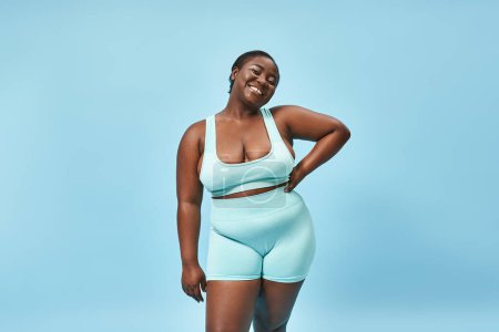 happy plus size sportswoman in biking shorts posing with hand on hip on blue backdrop, body positive