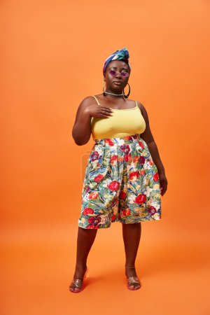 Foto de Elegante mujer afroamericana de talla grande con falda floral y pañuelo en la cabeza posando sobre fondo naranja - Imagen libre de derechos