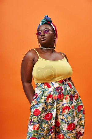 Foto de Elegante mujer afroamericana de talla grande con falda floral y gafas de sol posando sobre fondo naranja - Imagen libre de derechos