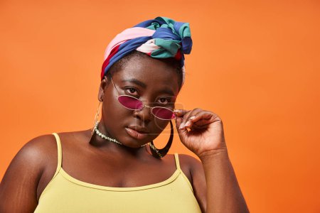 Fashion-forward and plus size african american model in headscarf and trendy sunglasses on orange