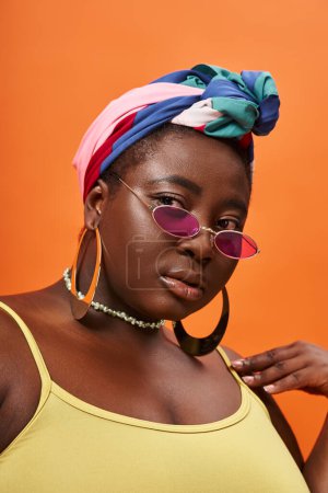 Photo pour Portrait de jeune femme afro-américaine taille plus en foulard et lunettes de soleil tendance sur orange - image libre de droit