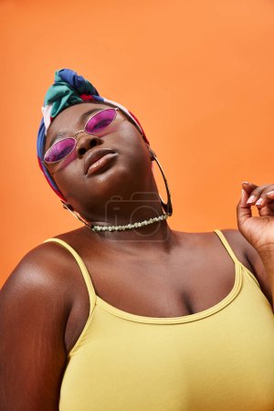 portrait of stylish plus size african american woman in headscarf and trendy sunglasses on orange