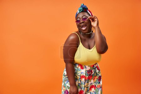 Foto de Mujer afroamericana feliz más tamaño con falda floral y gafas de sol posando sobre fondo naranja - Imagen libre de derechos