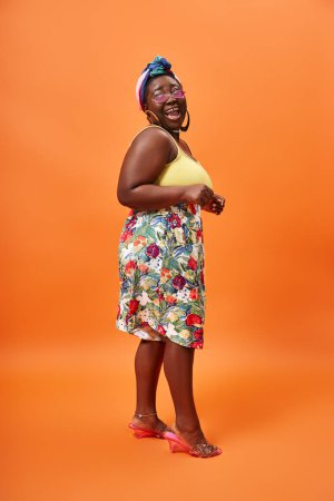 Foto de Sorprendido afroamericano más tamaño mujer con falda floral y gafas de sol posando sobre fondo naranja - Imagen libre de derechos