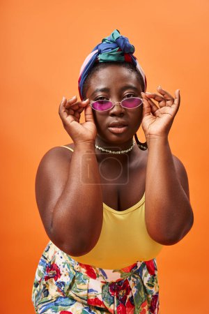 Photo pour Femme afro-américaine à la mode plus la taille en foulard portant des lunettes de soleil tendance sur orange - image libre de droit