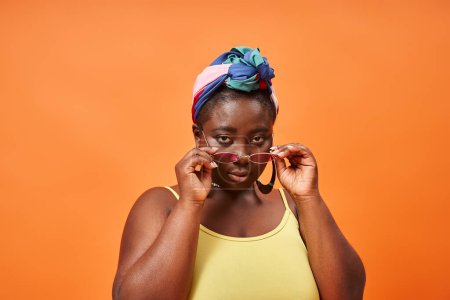 young plus size african american woman in headscarf and trendy sunglasses on orange backdrop