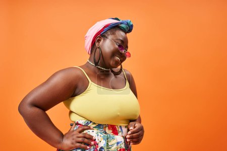 plus size african american woman in floral outfit and sunglasses posing with hand on hip on orange