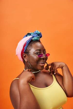 joyful plus size african american woman in headscarf and stylish sunglasses on orange backdrop