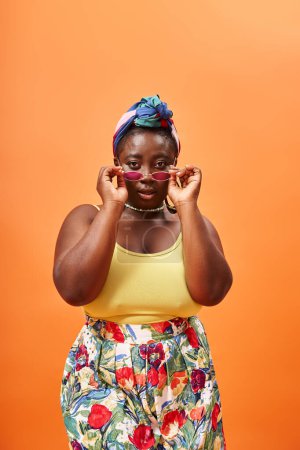 plus size african american woman in floral outfit wearing stylish sunglasses on orange background