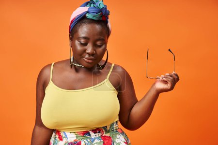 plus size african american woman in floral outfit holding trendy sunglasses on orange background
