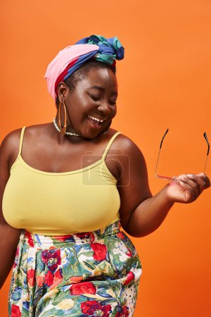 smiling plus size african american woman in floral outfit holding sunglasses on orange backdrop