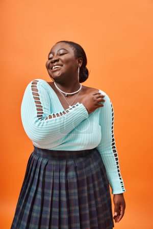 Photo for Cheerful plus size african american woman in plaid skirt and blue long sleeve on orange background - Royalty Free Image