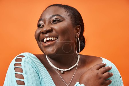 Téléchargez les photos : Portrait de femme afro-américaine gaie plus la taille en bleu manches longues sur fond orange - en image libre de droit