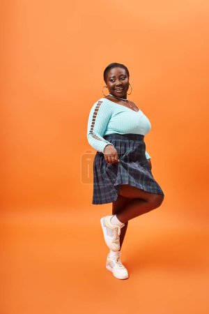 Photo for Happy plus size african american woman in plaid skirt and blue long sleeve on orange background - Royalty Free Image