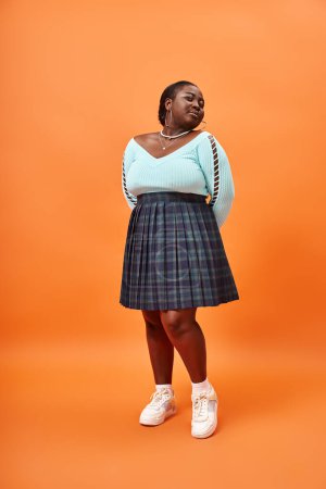 Téléchargez les photos : Jeune femme afro-américaine taille plus en jupe à carreaux et bleu manches longues sur fond orange - en image libre de droit