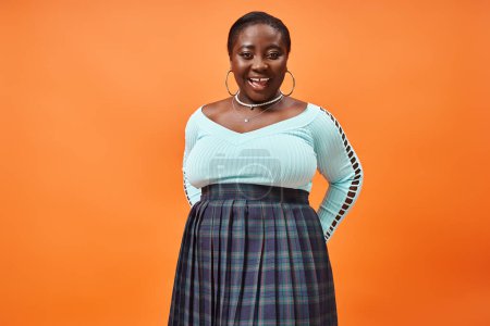 Téléchargez les photos : Corps positif femme afro-américaine en jupe à carreaux et bleu manches longues souriant sur fond orange - en image libre de droit