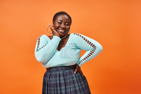 Téléchargez les photos : Corps positif femme afro-américaine en jupe à carreaux et bleu manches longues souriant sur orange - en image libre de droit