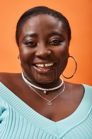 Téléchargez les photos : Portrait de corps positif jeune femme afro-américaine en bleu manches longues sur fond orange - en image libre de droit