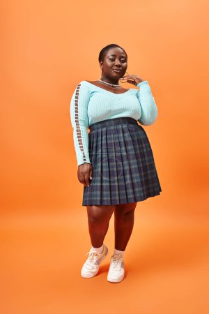Téléchargez les photos : Modèle afro-américain plus la taille en jupe à carreaux et pullover couleur menthe posant sur fond orange - en image libre de droit