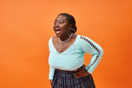 Photo for Amazed plus size african american model in plaid skirt and blue long sleeve posing on orange - Royalty Free Image