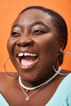 portrait of radiant plus size african american woman in blue long sleeve laughing on orange