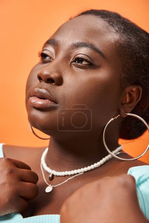 portrait of pensive plus size african american woman in blue long sleeve posing on orange backdrop