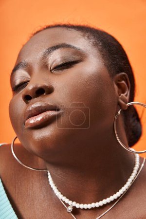 Porträt einer nachdenklichen plus size afrikanisch-amerikanischen Frau, die mit geschlossenen Augen auf orangefarbenem Hintergrund posiert