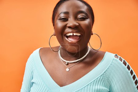 portrait of cheerful plus size african american woman in blue long sleeve laughing on orange