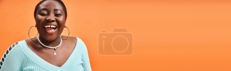Téléchargez les photos : Portrait de femme afro-américaine gaie et de taille plus en bleu manches longues riant sur orange, bannière - en image libre de droit