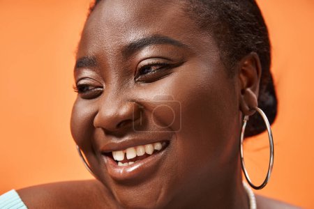 Téléchargez les photos : Portrait de femme afro-américaine gaie et de taille plus en bleu manches longues souriant sur orange - en image libre de droit