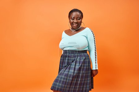 Téléchargez les photos : Gai plus la taille femme afro-américaine en jupe à carreaux et manches longues souriant sur fond orange - en image libre de droit