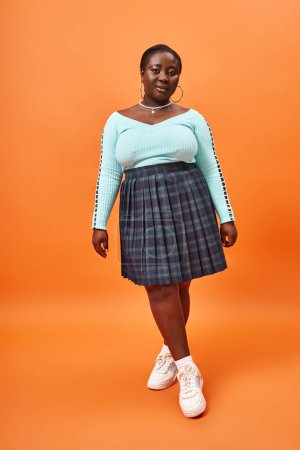 Téléchargez les photos : Pleine longueur, plus la taille femme afro-américaine en jupe à carreaux et manches longues posant sur orange - en image libre de droit