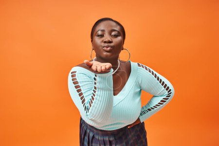 Téléchargez les photos : Femme afro-américaine taille plus en jupe à carreaux et manches longues envoyer un baiser d'air sur orange - en image libre de droit