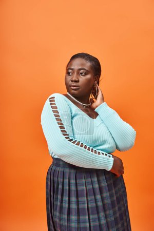 portrait of charming plus size african american woman in blue long sleeve and plaid skirt on orange