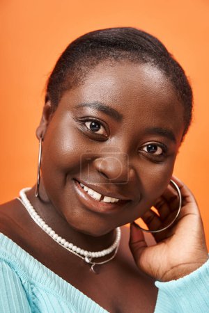 portrait of joyful plus size african american woman in blue long sleeve smiling on orange