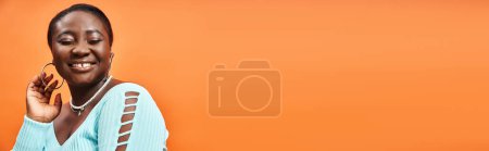 portrait de femme africaine américaine gaie et de taille plus en bleu manches longues souriant sur orange, bannière