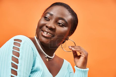 portrait of pleased plus size african american woman touching her hoop earring and smiling on orange