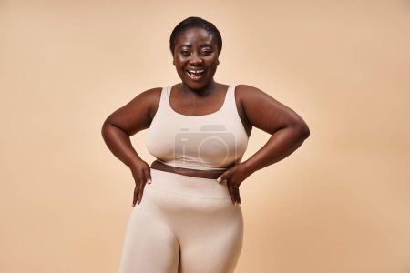 Radiante mujer afroamericana de tamaño grande en ropa deportiva beige con sonrisa alegre en el fondo a juego