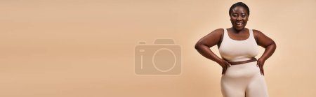 Radiant plus size african american woman in beige sportswear smiling on matching backdrop, banner
