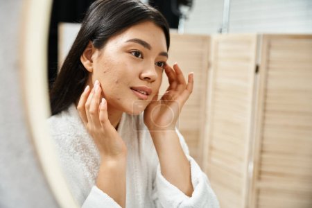 junge asiatische Frau mit brünetten Haaren untersucht ihre Haut im Badezimmerspiegel, Hautzustand
