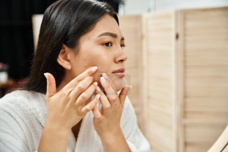 junge asiatische Frau mit brünetten Haaren untersucht ihre Haut mit Akne im Badezimmerspiegel, Hautproblem